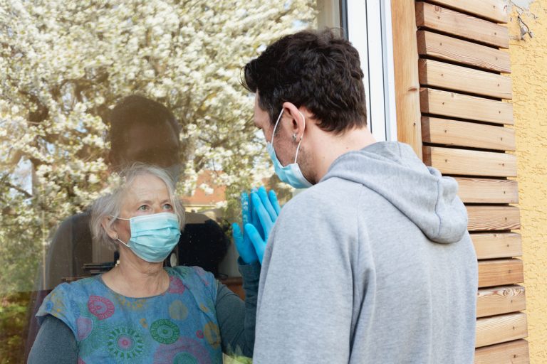 Senior woman wearing mask visiting middle age man wearing mask through window