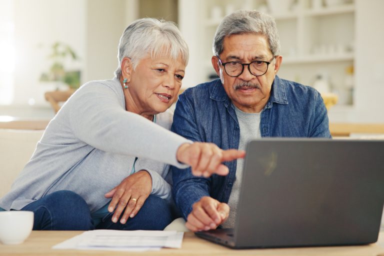 Elderly Hispanic couple, woman on the left and man on the right, look at a laptop on their coffee table. Woman has short white hair and a blue sweater. Man has dark gray hair, a white t-shirt, and a blue flannel on. The woman is pointing to the laptop.